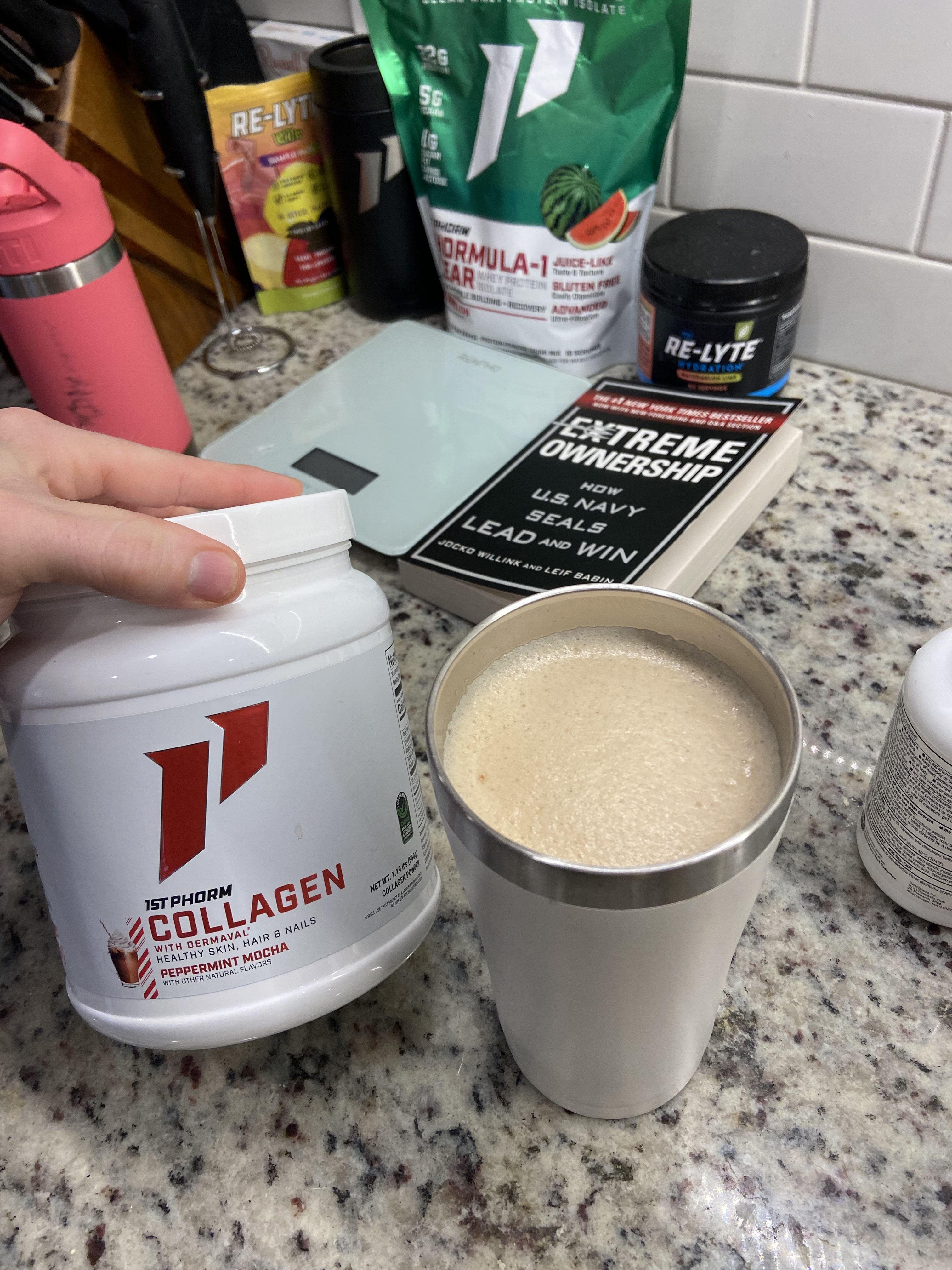 Hand holding 1st Phorm Collagen Container next to a mug of steaming hot coffee